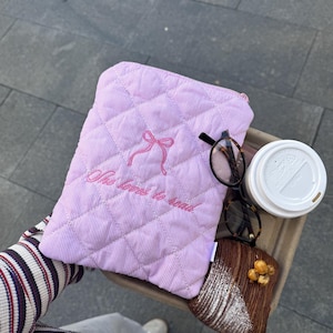 May include: A pale pink, quilted book sleeve with the embroidered text "She loves to read" and a bow design. Tortoiseshell glasses, a croissant, and a coffee cup are on a tray.