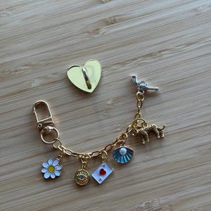 May include: A gold-coloured keychain with a lobster clasp and a chain holding various charms. Charms include a white daisy, an eye, a playing card, a seashell, a leopard, and a small revolver. A gold heart-shaped pin is also present.