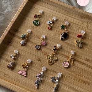 Puede incluir: Una colección de pendientes con colgantes variados: fresa, mariposa, carta de juego, cereza, trébol de cuatro hojas, balón de fútbol americano, lazo, tortuga, sol, flor, oso de peluche, sombrero de vaquero, dragón, mariposa, cruz, libélula y espada.