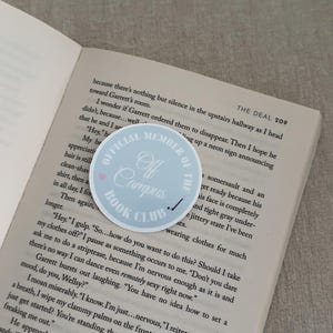 May include: A light blue sticker with the text "Official Member of the Off Campus Book Club" and a small pink heart, placed on an open book. The book's pages are visible, with text from a novel.