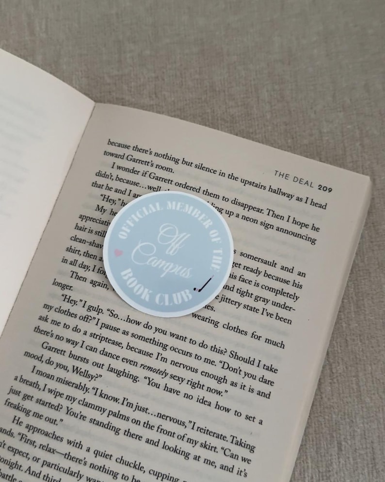 May include: A light blue sticker with the text "Official Member of the Off Campus Book Club" and a small pink heart, placed on an open book. The book's pages are visible, with text from a novel.