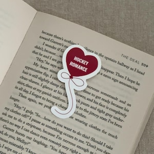 May include: A heart-shaped sticker with the words "HOCKEY ROMANCE" in white on a red background. The sticker is on a white background and has a balloon-like design. The sticker is placed on an open book.