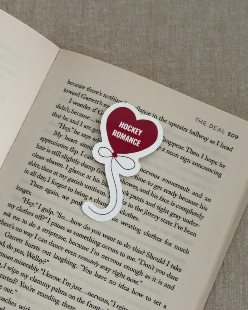 May include: A heart-shaped sticker with the words "HOCKEY ROMANCE" in white on a red background. The sticker is on a white background and has a balloon-like design. The sticker is placed on an open book.