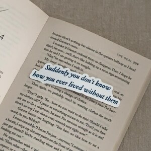 May include: A white bookmark with the text "Suddenly you don't know how you ever lived without them" in a dark blue font, placed on an open book. The book's pages are light beige.