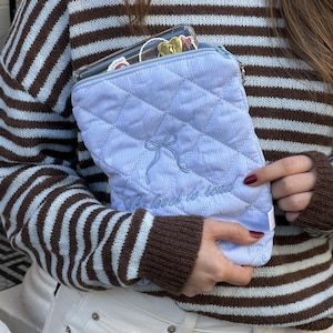 Könnte beinhalten: Eine hellblaue, gesteppte Tasche mit Schleifenmuster und dem gestickten Text "Whatever it takes". Die Tasche wird von einer Person gehalten, die einen braun-hellblau gestreiften Pullover und eine weiße Jeans trägt. Eine transparente Handyhülle ist in der Tasche sichtbar.