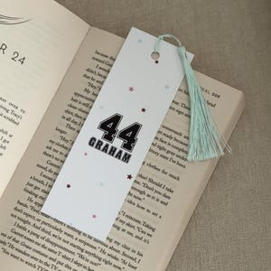 May include: White bookmark with the number "44" and the name "GRAHAM" in black. The bookmark has a light blue tassel and small star accents. It is placed in an open book.