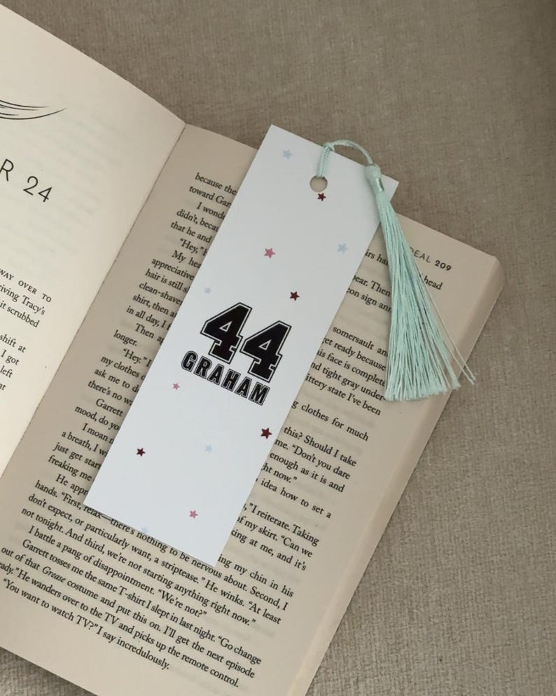 May include: White bookmark with the number "44" and the name "GRAHAM" in black. The bookmark has a light blue tassel and small star accents. It is placed in an open book.