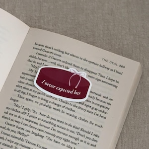 May include: A burgundy-colored sticker with the text "I never expected her" in white, featuring a white bow design. The sticker is placed on an open book with printed text.