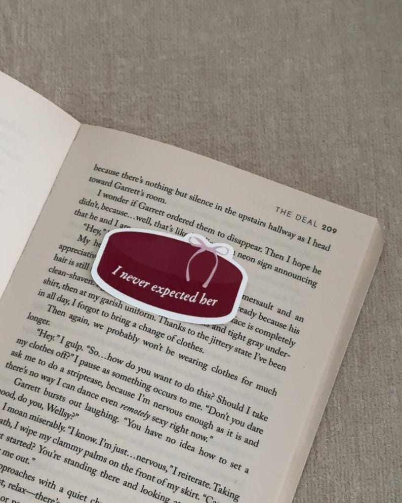 May include: A burgundy-colored sticker with the text "I never expected her" in white, featuring a white bow design. The sticker is placed on an open book with printed text.