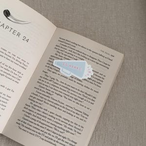 May include: A light blue and white "Go Hawks" megaphone sticker on an open book. The book is open to Chapter 24, with text on the pages. The sticker has a pink burst design.