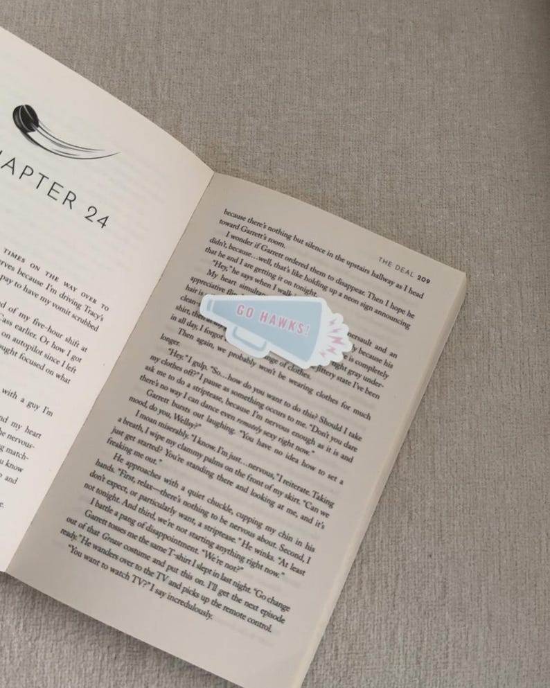 May include: A light blue and white "Go Hawks" megaphone sticker on an open book. The book is open to Chapter 24, with text on the pages. The sticker has a pink burst design.
