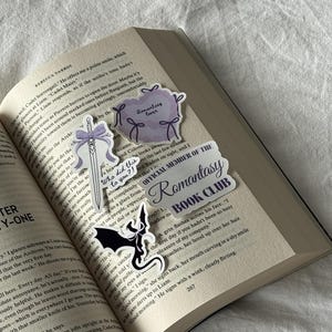 May include: An open book with several stickers on the pages. The stickers include a purple cloud with the text "Romantasy lover", a sword with a purple bow, a sticker with a black bat, and a sticker that says "OFFICIAL MEMBER OF THE Romantasy BOOK CLUB".