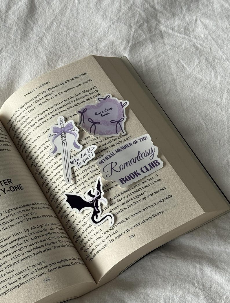 May include: An open book with several stickers on the pages. The stickers include a purple cloud with the text "Romantasy lover", a sword with a purple bow, a sticker with a black bat, and a sticker that says "OFFICIAL MEMBER OF THE Romantasy BOOK CLUB".