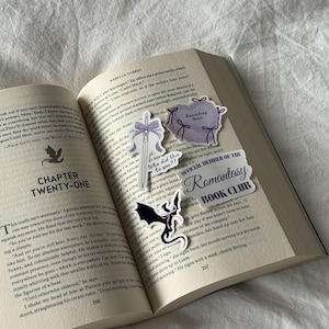 May include: An open book with several decorative stickers. The stickers include a sword with a purple bow, a purple heart with bows, a black dragon, and a banner that reads "Romantasy Book Club."