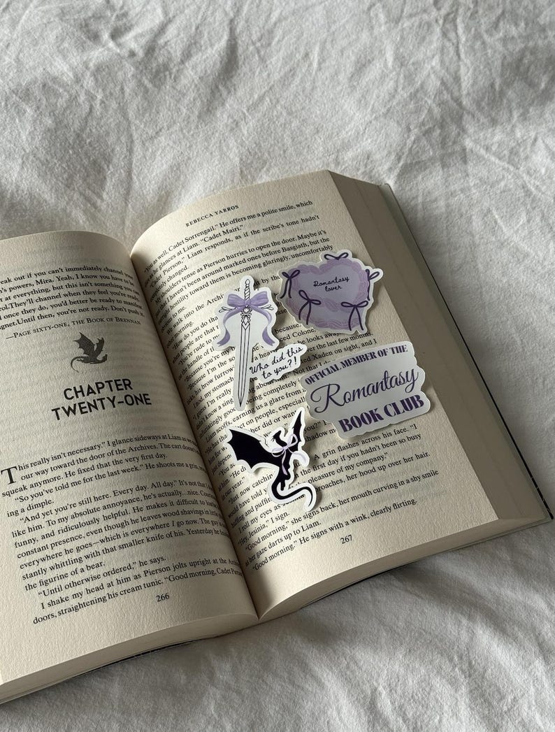 May include: An open book with several decorative stickers. The stickers include a sword with a purple bow, a purple heart with bows, a black dragon, and a banner that reads "Romantasy Book Club."