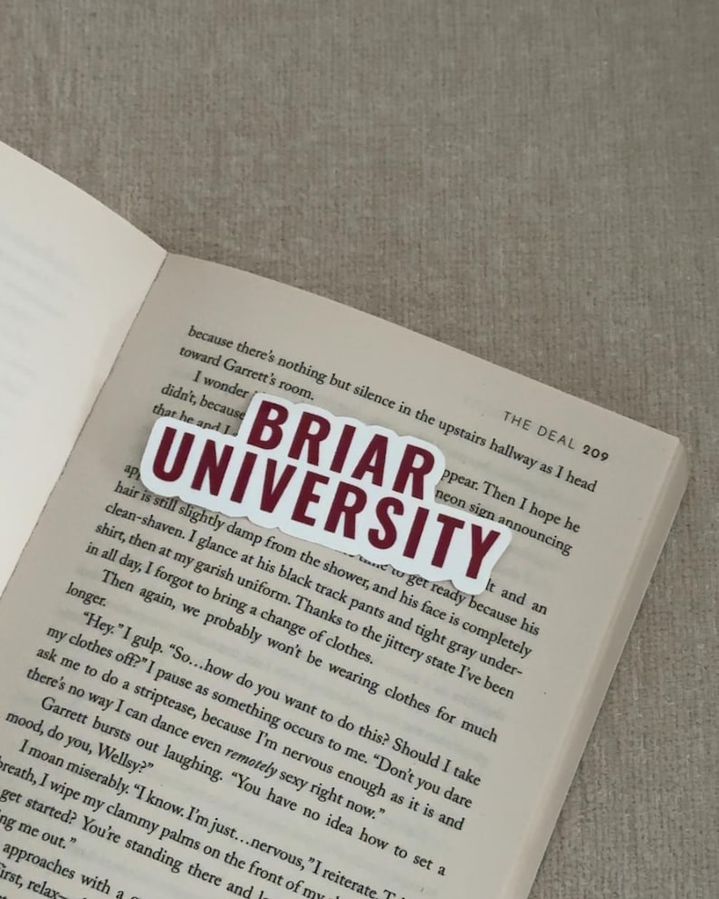 May include: A white sticker with the text "BRIAR UNIVERSITY" in maroon letters is placed on an open book. The book's pages are filled with printed text. The sticker is rectangular with a white border.