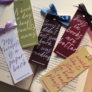 May include: Five rectangular paper bookmarks with ribbons, each featuring unique text: 'My Therapist is a paperback', 'A book a day keeps reality away', 'Sorry I didn't text you back, I was reading', 'Boys in books are better', and 'I would like to stay in bed & read please'.