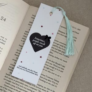 May include: White bookmark with a black heart design and the text "Sometimes, people sneak up on you." The bookmark has a light blue tassel and small star accents. It is placed on an open book.