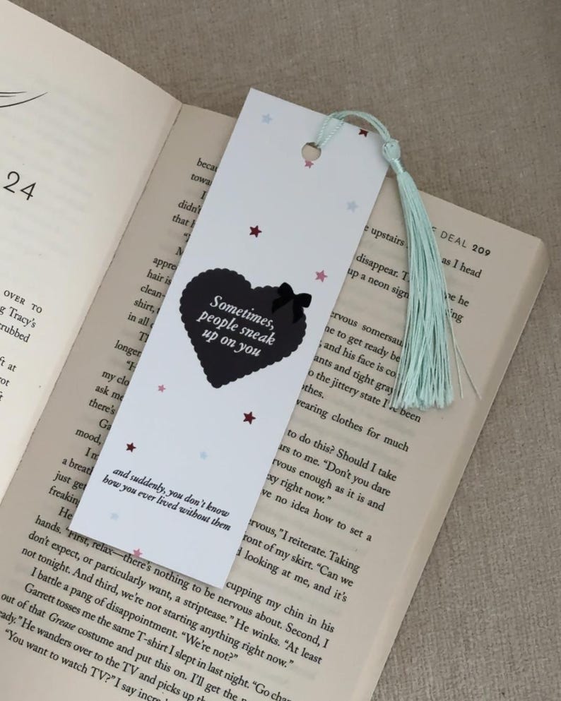 May include: White bookmark with a black heart design and the text "Sometimes, people sneak up on you." The bookmark has a light blue tassel and small star accents. It is placed on an open book.