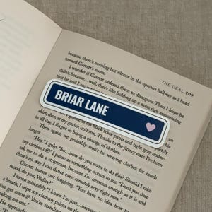 May include: A rectangular bookmark with the text "BRIAR LANE" in white on a navy blue background, with a small pink heart. The bookmark is placed on the open pages of a book.
