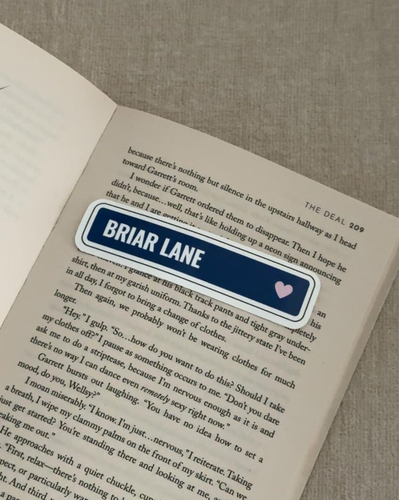May include: A rectangular bookmark with the text "BRIAR LANE" in white on a navy blue background, with a small pink heart. The bookmark is placed on the open pages of a book.