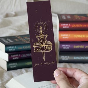 Peut inclure: Un marque-page bordeaux avec des détails dorés, représentant une épée traversant des livres, avec l'inscription "to whatever end, you do not yield". Le marque-page est devant une pile de livres.
