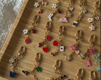 Op de afbeelding: Een verzameling goudkleurige sleutelhangers met diverse bedels, waaronder bloemen, harten, aardbeien en een koffiekopje. De bedels zijn in roze, rood en blauw, gerangschikt op een houten dienblad.