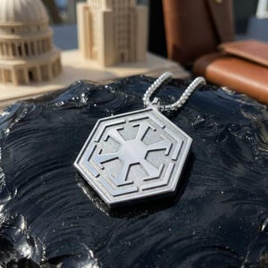 Peut inclure: Un pendentif hexagonal argenté avec une chaîne. Le pendentif présente un motif symétrique détaillé avec une étoile centrale et des motifs géométriques. Le collier est présenté sur une surface sombre et texturée.