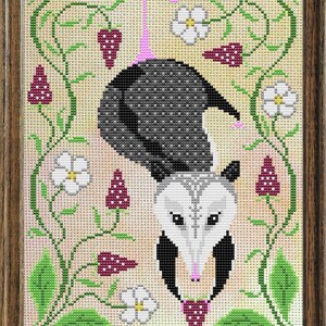 Könnte beinhalten: Kreuzstich-Kunstwerk eines kopfüber hängenden Opossums, umgeben von weißen Blumen, grünen Ranken und lila Beeren. Das Opossum hat einen schwarz-grauen Körper, ein weißes Gesicht und eine rosa Nase. Das Kunstwerk ist in einem Holzrahmen gerahmt.