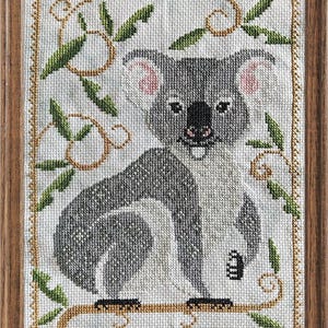 May include: A framed cross-stitch embroidery featuring a grey and white koala sitting on a branch. The koala has pink inner ears and a black nose. The design is surrounded by a decorative border of golden-brown vines and green leaves on white fabric.