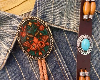 May include: Oval bolo tie with a detailed moth design in shades of orange, teal, and red, set against a dark background. The tie features a braided leather cord and a brown leather strap with a turquoise stone accent. The text "CowboyHat wild life" is visible.