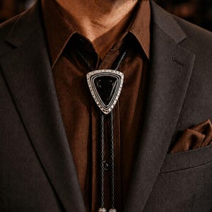 May include: A triangular bolo tie with a black stone center and a silver-colored border. The tie is worn with a brown button-down shirt and a dark gray suit jacket. A brown pocket square is visible.