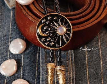 Op de afbeelding: Een zwarte en gouden bolo stropdas met een hemels ontwerp met een maan, zon en sterren. De stropdas heeft een gevlochten zwart koord en decoratieve gouden uiteinden. De afbeelding toont ook een bruine leren riem en verschillende witte parels.