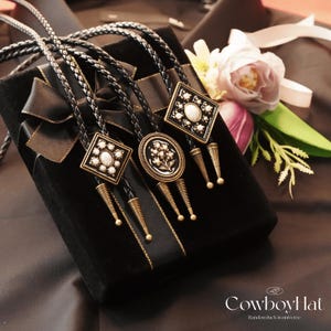 May include: Three bolo ties with decorative square and oval pendants, featuring white stones and floral designs, rest on a black velvet box with a black ribbon. The ties have braided leather cords and gold-colored metal accents. A flower and greenery are in the background.