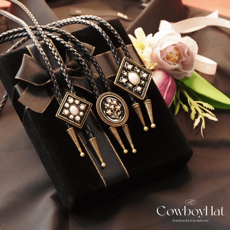 May include: Three bolo ties with decorative square and oval pendants, featuring white stones and floral designs, rest on a black velvet box with a black ribbon. The ties have braided leather cords and gold-colored metal accents. A flower and greenery are in the background.