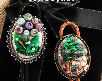 May include: Two embroidered bolo ties. One features a floral design with pink, green, and purple accents, and the other showcases a Christmas tree with gifts. Both have decorative metal frames and leather cords. The word "Cowboyhat" is at the top.