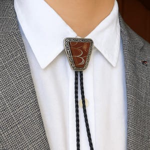 May include: A bolo tie with a triangular pendant featuring a brown and cream-coloured design. The pendant is framed by a silver-coloured, ornate border. The tie has a black braided cord and is worn with a white collared shirt and a grey blazer.