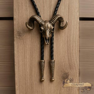 May include: A bronze-colored ram skull bolo tie with a black braided leather cord. The skull has detailed horns and is centered on the cord. The cord ends with decorative bronze-colored tips. The tie is displayed on a wooden surface.