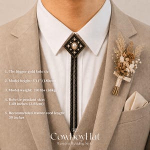 May include: A gold bolo tie with a diamond-shaped pendant featuring a central pearl and surrounding embellishments. The black leather cord hangs over a white collared shirt, paired with a beige suit. A small floral boutonniere and the text "Cowboy Hat" are also visible.