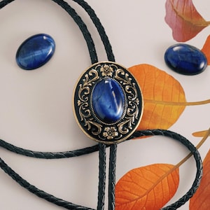 May include: A bolo tie with a large oval blue stone center, surrounded by an ornate gold and black filigree design. Two matching blue oval stones are also present. The tie features a black braided cord.