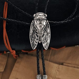 May include: A silver cicada bolo tie with a detailed insect design. The bolo tie has a braided black leather cord and decorative silver tips. The background includes a black leather surface and tan fabric.