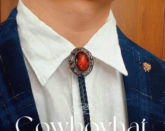 May include: A bolo tie with a dark red oval stone set in an ornate, bronze-colored frame. The tie features a braided navy blue cord and is worn over a white shirt and a blue plaid blazer. The text "Cowboyhat" is at the bottom.