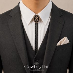 May include: A black and gold bolo tie with a floral design is the focal point of this image. The tie is worn with a white collared shirt, a dark gray suit jacket, and a matching vest. A cream-colored pocket square adds a touch of elegance. The text "CowboyHat Western Wedding Style" is at the bottom.