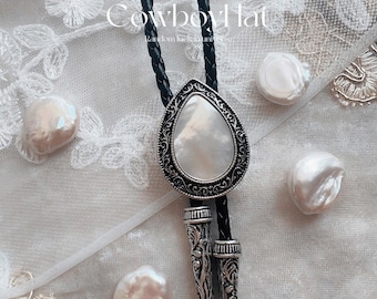 May include: A bolo tie with a pearlescent, teardrop-shaped pendant, framed by an ornate silver-toned border. The black braided cord is visible, along with decorative silver tips. The text "Cowboy Hat" and "Suitable for weddings, cowboy style, and vintage-inspired looks" are also visible.