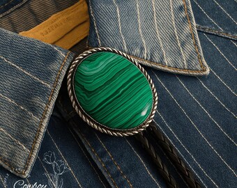 May include: Oval malachite bolo tie with a silver-toned rope-like border. The green stone has dark green veining. The tie is on a blue denim shirt with white pinstripes. The bolo tie has black leather cords and a white button.