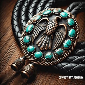 Puede incluir: Una corbata bolo con un colgante de águila de bronce, adornado con piedras de color turquesa. El colgante está engastado sobre un cordón de cuero trenzado negro. El texto "COWBOY HAT JEWELRY" es visible en la parte inferior.