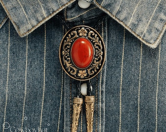 May include: A bolo tie with a large oval red stone set in an ornate, gold-colored metal frame. The tie features a black cord and decorative gold-colored tips, set against a blue and white striped denim shirt.