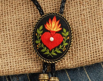 May include: Oval bolo tie with a black background featuring a red heart with a flame and pearl accent, surrounded by green leaves. The tie has a braided black cord and decorative gold-colored tips.