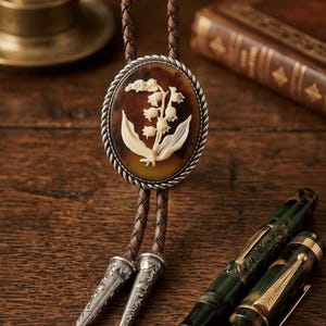 Puede incluir: Una corbata bolo con un cordón de cuero marrón y un colgante ovalado. El colgante presenta un diseño de lirio de los valles blanco sobre un fondo de carey, enmarcado por un borde plateado en forma de cuerda. También se ven dos plumas ornamentadas.