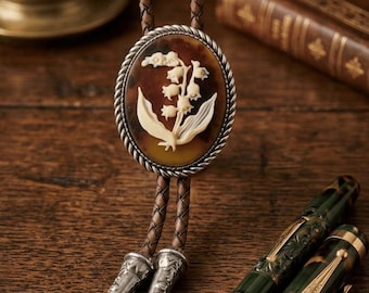 May include: A bolo tie with a brown leather cord and a decorative oval pendant. The pendant features a white lily of the valley design set against a tortoiseshell background, framed by a silver rope-like border. Two ornate pens are also visible.
